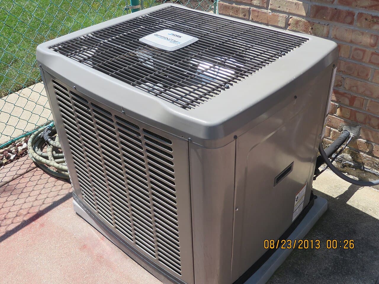 An outdoor central air conditioning condenser unit on a concrete pad beside a brick house