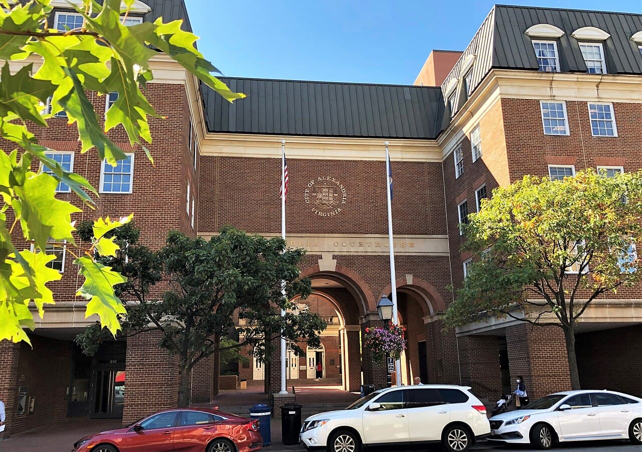 Red-brick Alexandria Courthouse on King Street with arched entrance, city seal, two flagpoles and cars parked in front