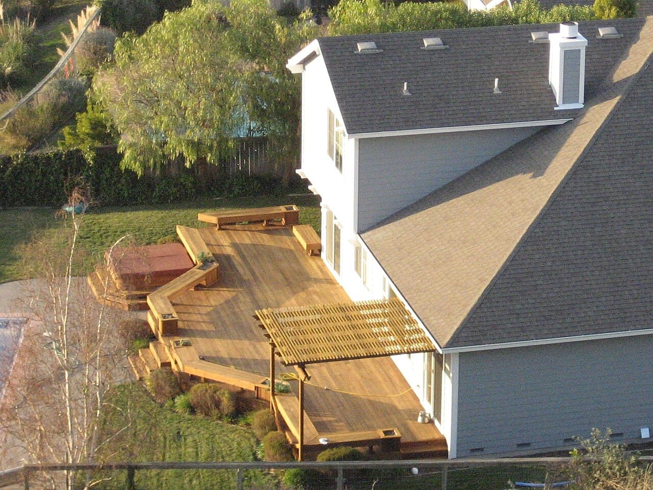 A large wooden deck with built-in benches, steps and a pergola, attached to the back of a grey two-story house