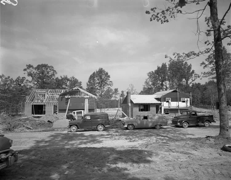 Black-and-white photo of brick houses being built, one still in its roof framing, with work trucks parked in front