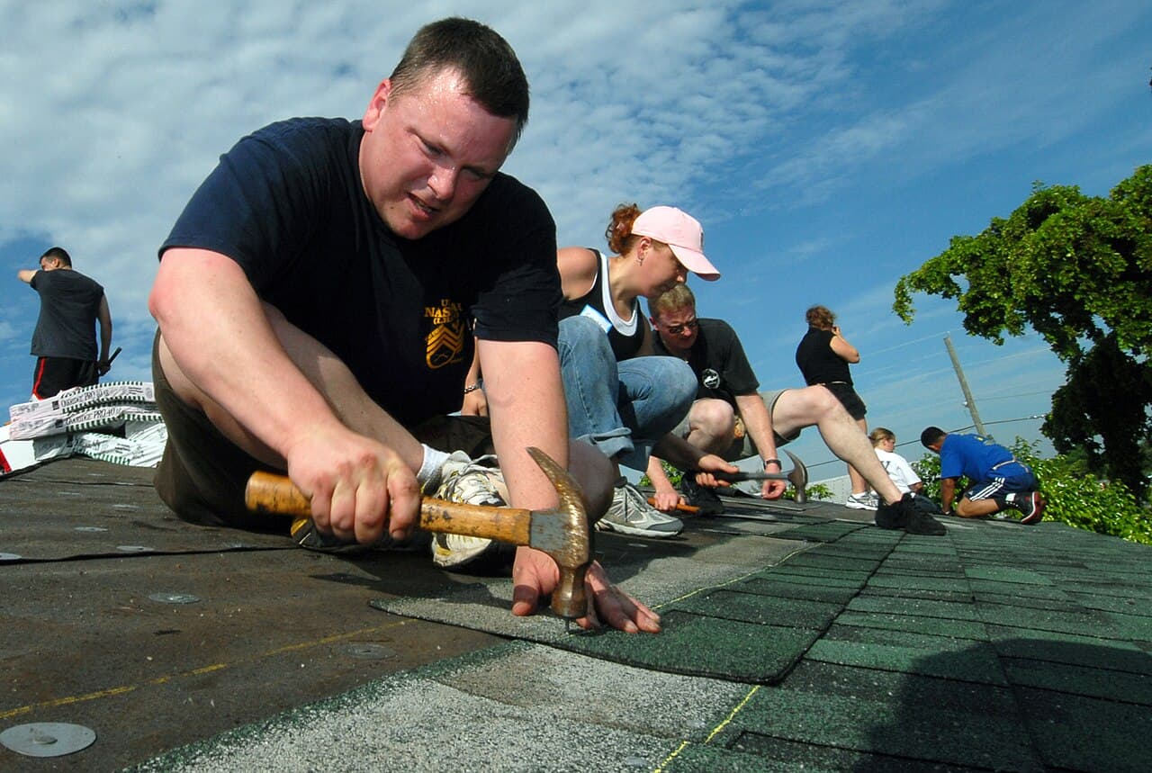 Volunteers kneel on a house roof, nailing new green asphalt shingles over the underlayment
